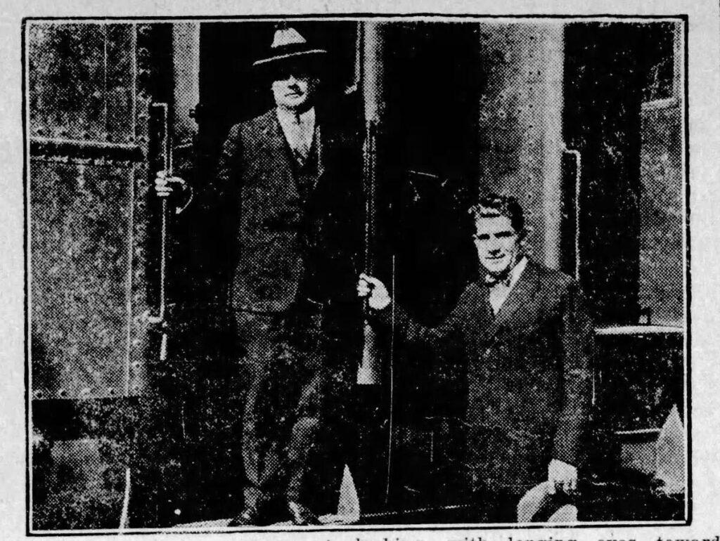 Black and white photograph of two men in suits, one wearing a fedora, standing in a doorway, possibly a train entrance, in a grainy newspaper clipping style.